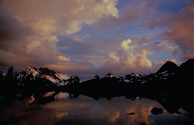 A mountain lake reflects a mountain peak with glaciers and dramatic, sunset colored clouds.