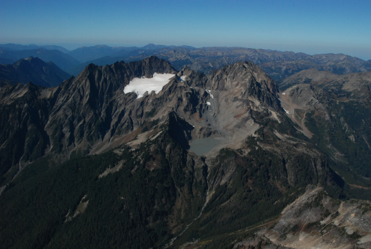 An aerial view of mountain peaks. One peak has a large glacier on one side, and a much smaller field of snow on another, draining to a lake basin.