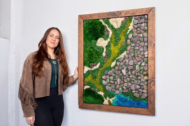 A woman in a fringed suede jacket stands beside a framed art piece. Within the wood frame, rocks and colored moss form the shapes of a landscape.