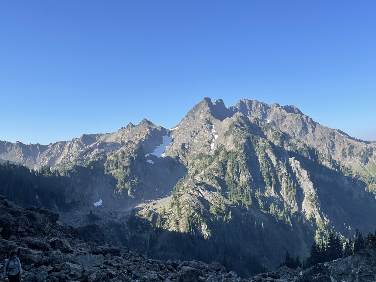 A mountain below a clear blue sky. A small glacier nestles in the crook between two peaks. A person wearing a backpack is visible in the foreground.