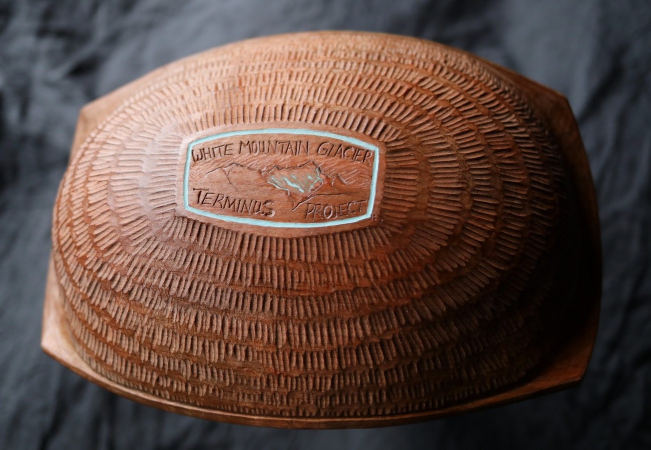 The bottom of a carved wooden bowl, etched in short lines forming concentric circles around a flat bottom with the words "White Mountain Glacier Terminus Project" and a small glacier in a mountainside carved.