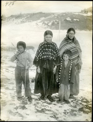 A Native American woman with three adolescent children standing in front of her. They are standing close together in a snowy open area looking at the photographer.