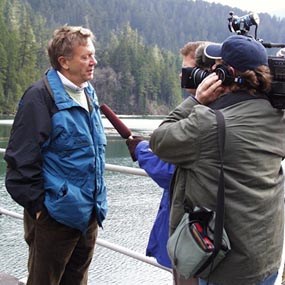 A man stands above a reservoir. The man is being interviewed by a news team consisting of an interviewer with a microphone and a videographer.