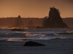 Waves break on a large beach cove. Rocky pinnacles known as "sea stacks" can be seen on the other side of the cove.