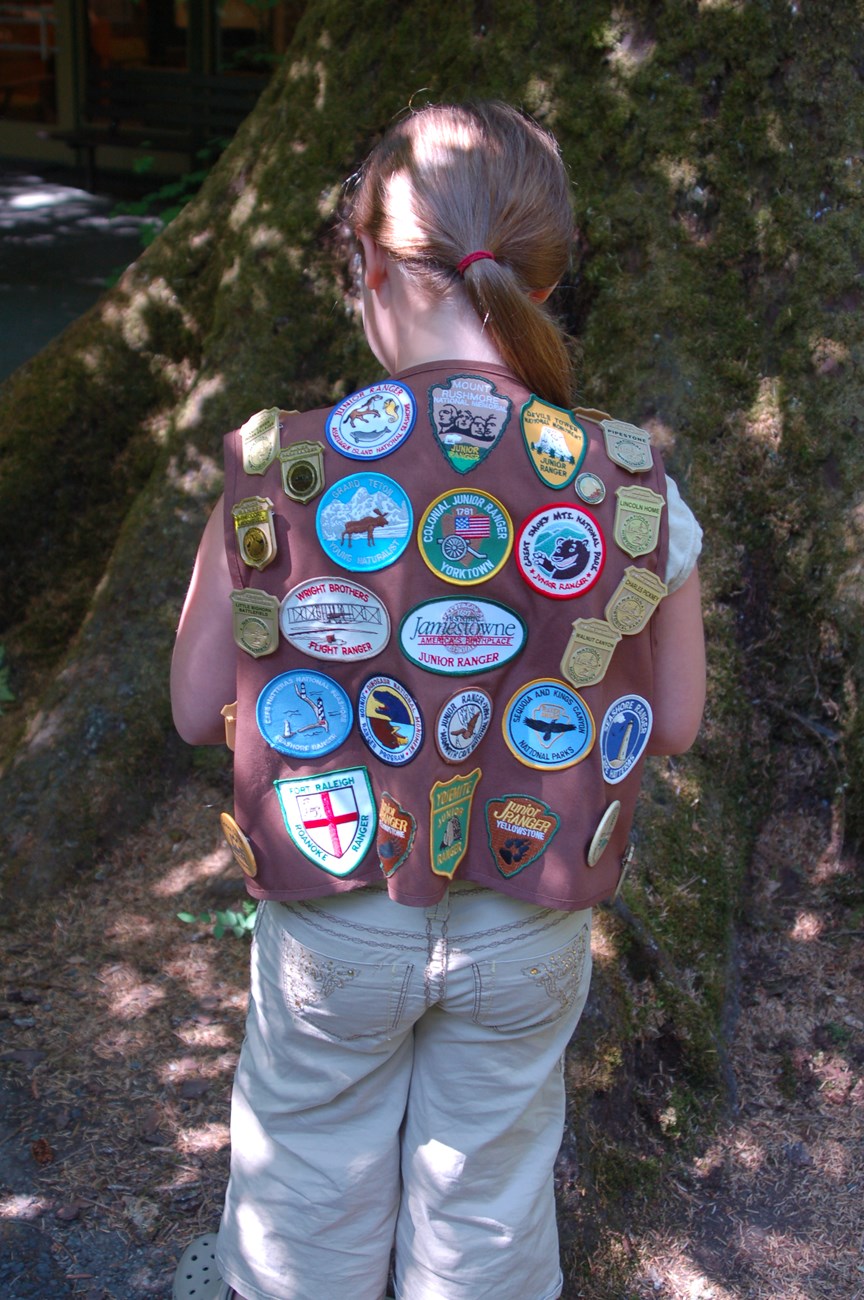 A young girl with a ponytail turned to show many junior ranger badges on the back of her vest.