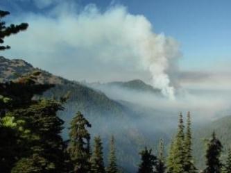 A large plume of smoke arises into the sky from a wildfire in a river valley. In addition to the plume, a smoky haze also sits over the river valley.