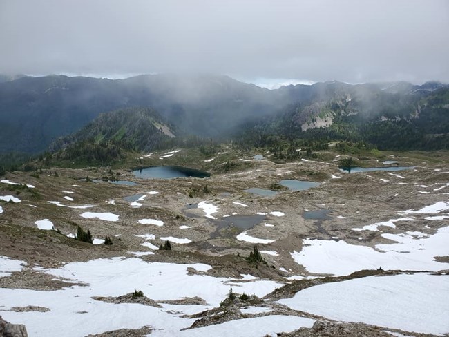 The image depicts alpine lakes in a rocky basin with clouds and a ridge behind them and snow on the ground around them.