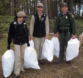 Two young volunteers stand next to a park Ranger near the coast. All three people each hold two bags of trash.