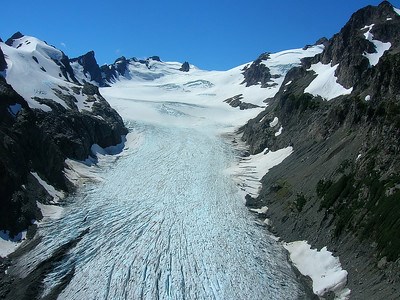 This image depicts the Hoh Glacier with a blue-ish white tint viewed from the terminus with grey rocky slopes on both sides and snow fields beyond it.