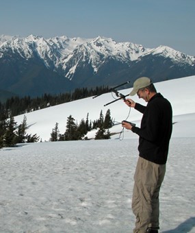 man holding small radio antenna on a snow-covered slope; snow-capped mountains in background