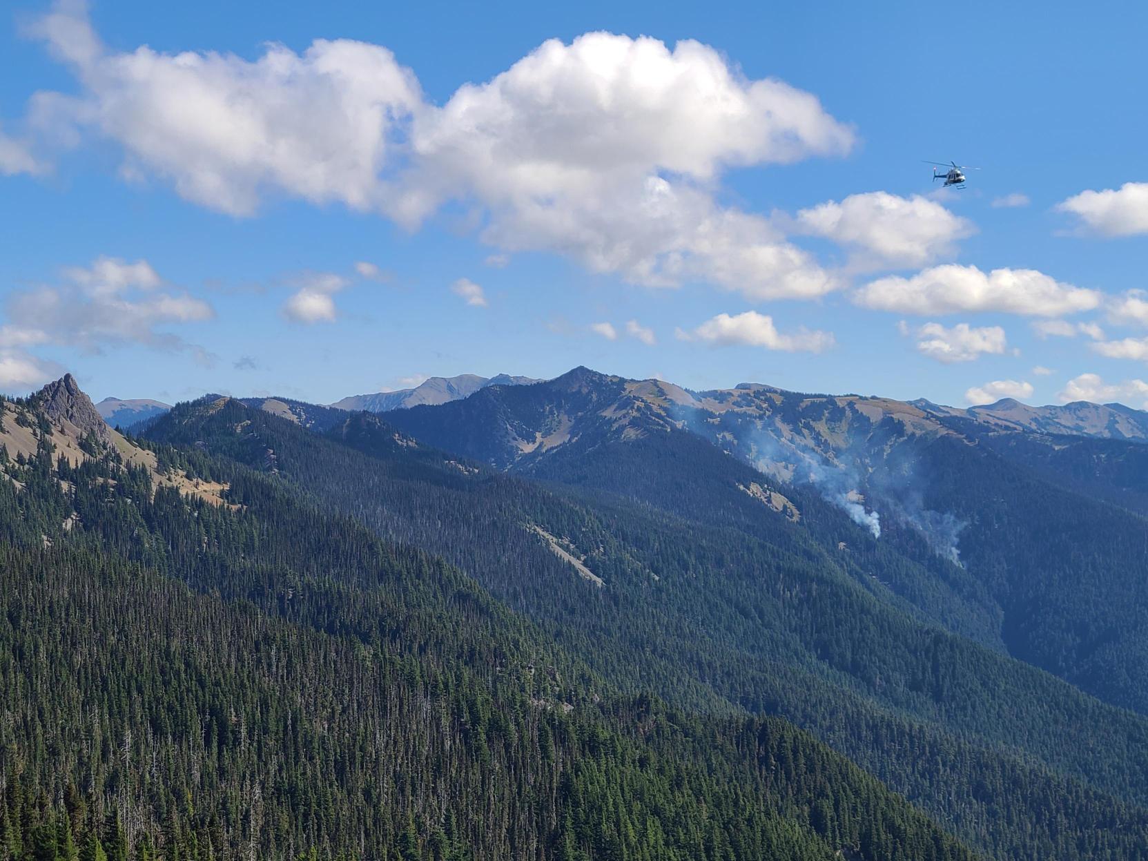 Eagle Point Fire with Helo Small smoke plumes can be seen above trees in the distance with a helicopter flying overhead.