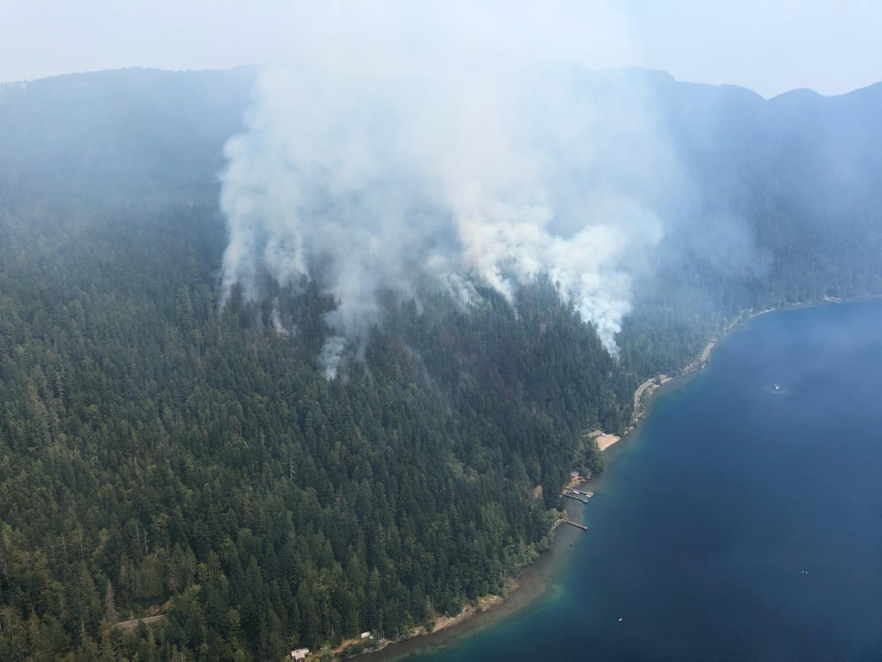An aerial photo of smoke from a wildfire burning in the forest next to a lake.
