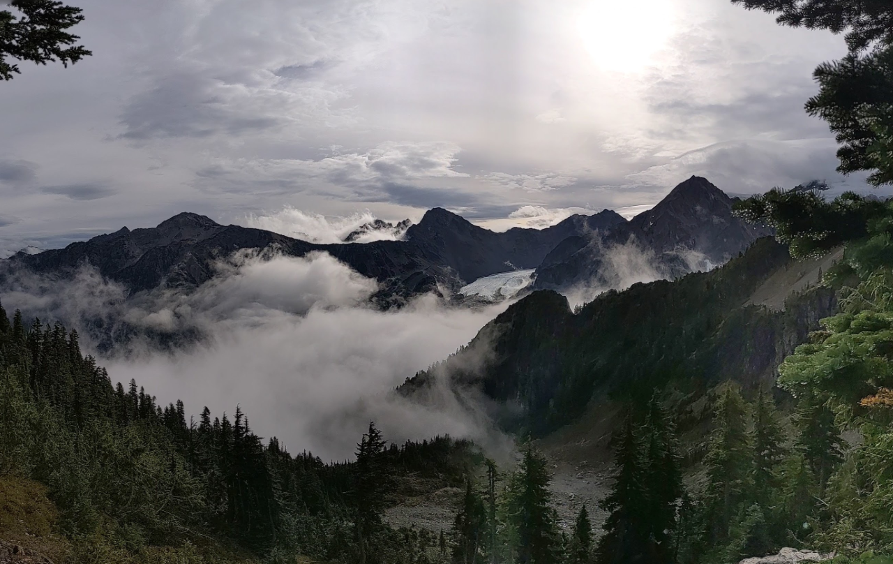 A mountainous, subalpine landscape with low clouds and a glacier in the distance emerging between two peaks