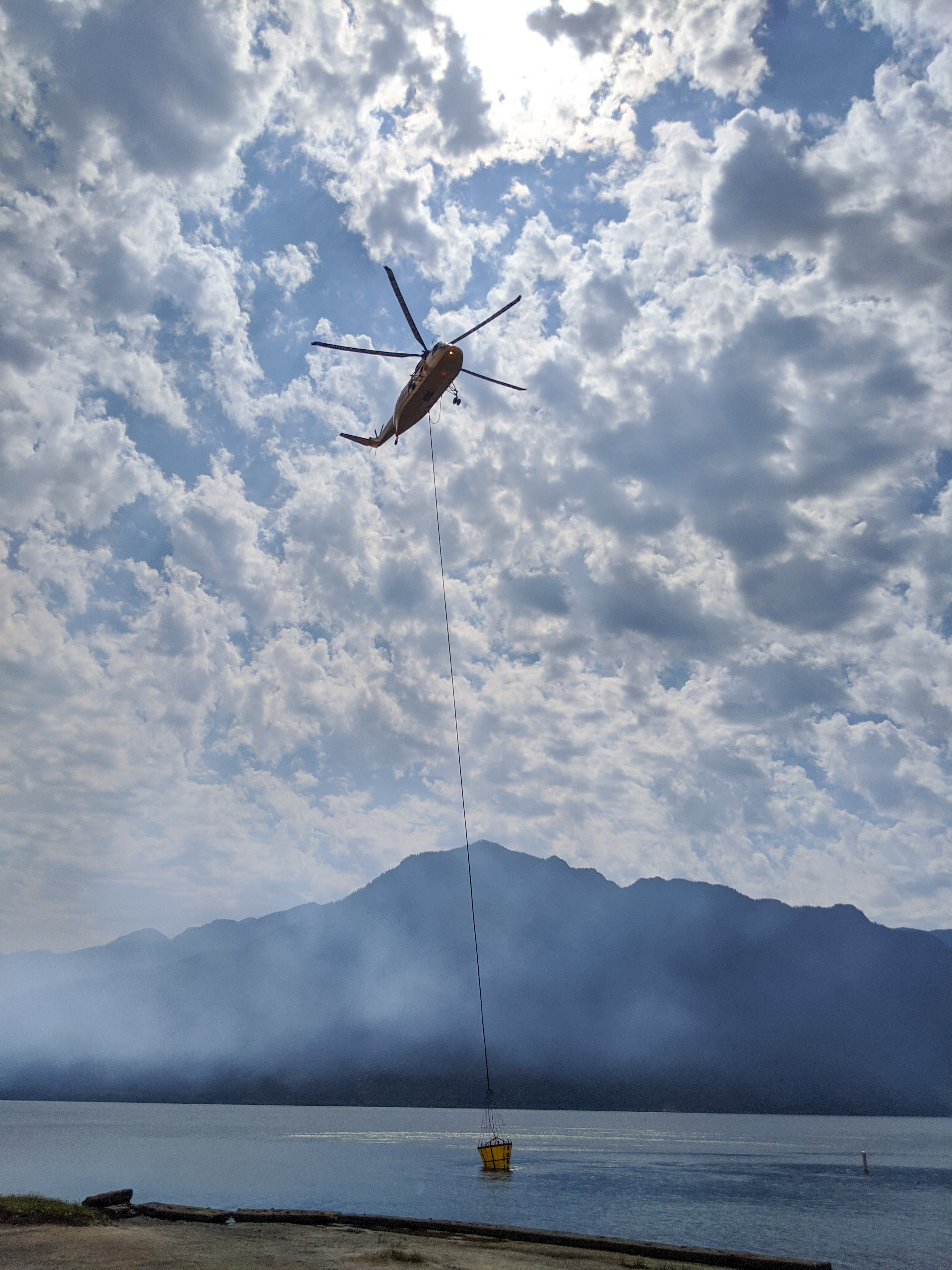 Helicopter filling a bucket with water from the lake to drop on the fire. A helicopter with a bucket suspended on a cable drawing water to drop on a nearby fire.
