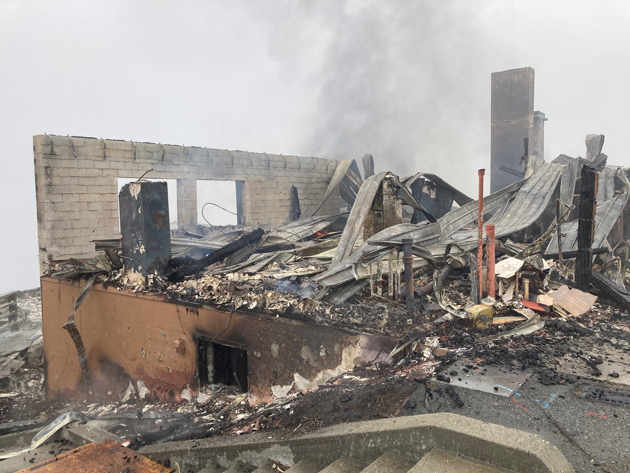 The burned remains of a building. Pipes stick up through the rubble and one cinderblock wall with openings that were once windows.