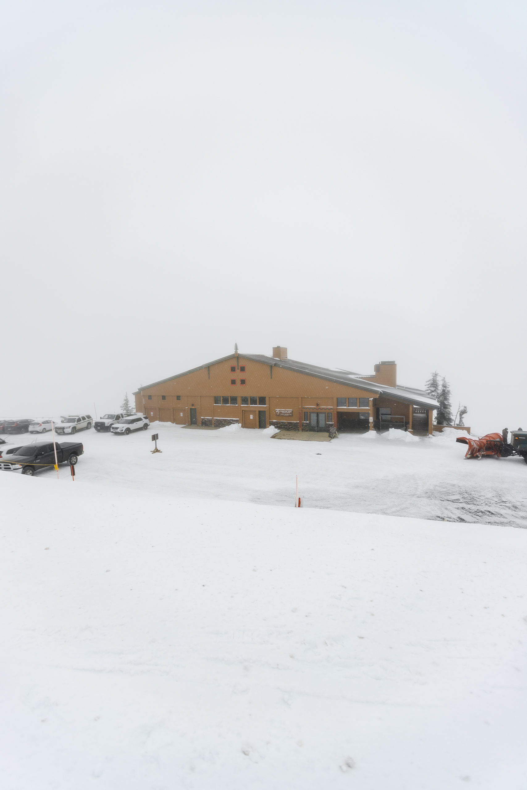 A wooden building in a snowy setting. Vehicles and a snowplow parked nearby.
