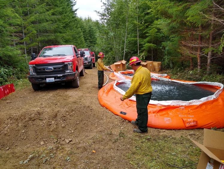 Firefighters are filling portable water tanks on top of the ridge above the fire. Two firefighters filling a portable water tank.