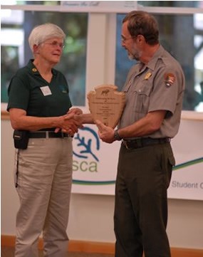 man in National Park Service uniform handing wooden arrowhead to woman wearing green polo shirt and khaki pants