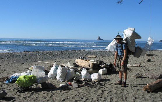 man with backpack standing near many filled garbage bags on ocean beach