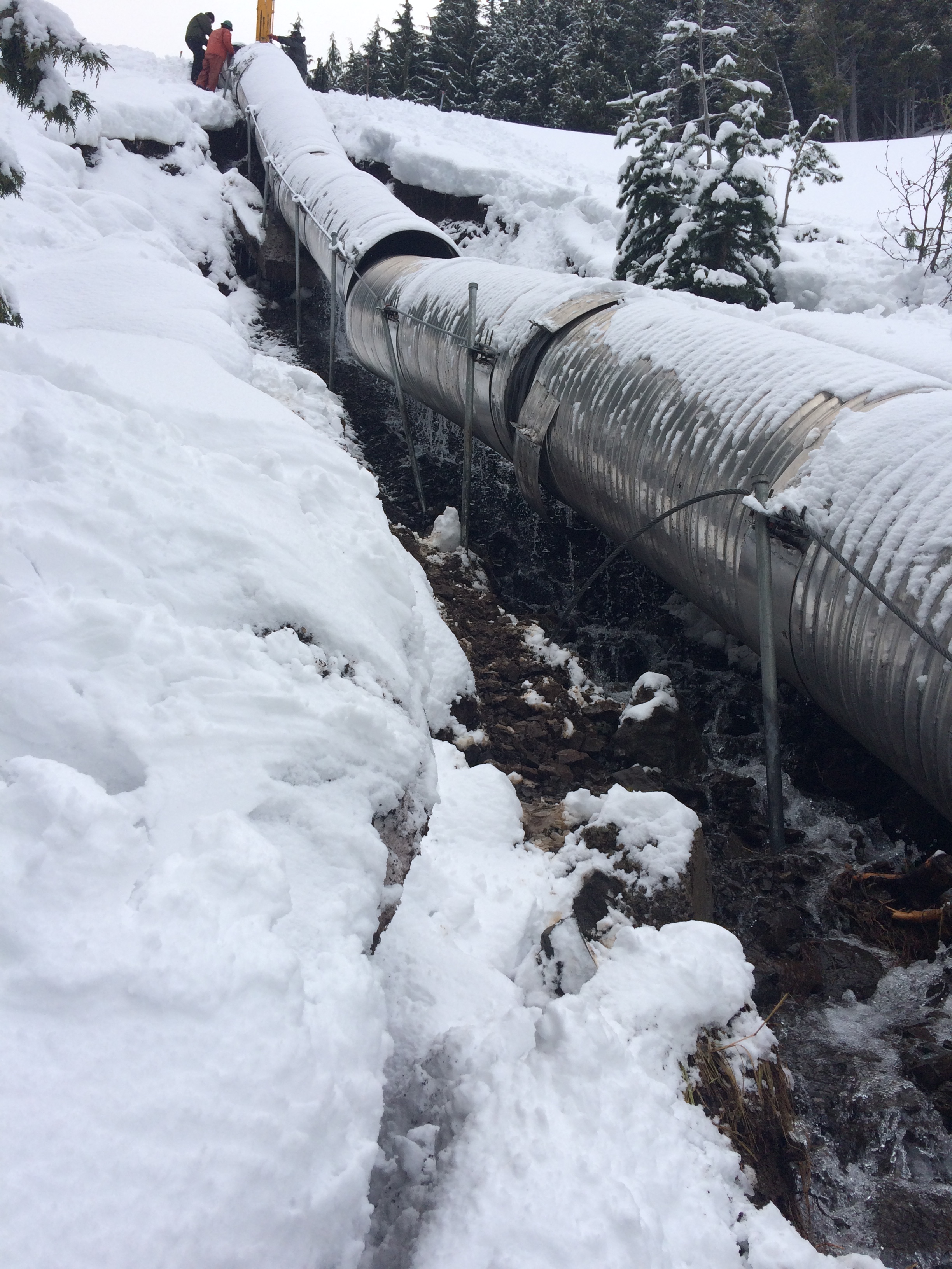 Broken culvert on snowy hillside with water draining underneath the pipe.