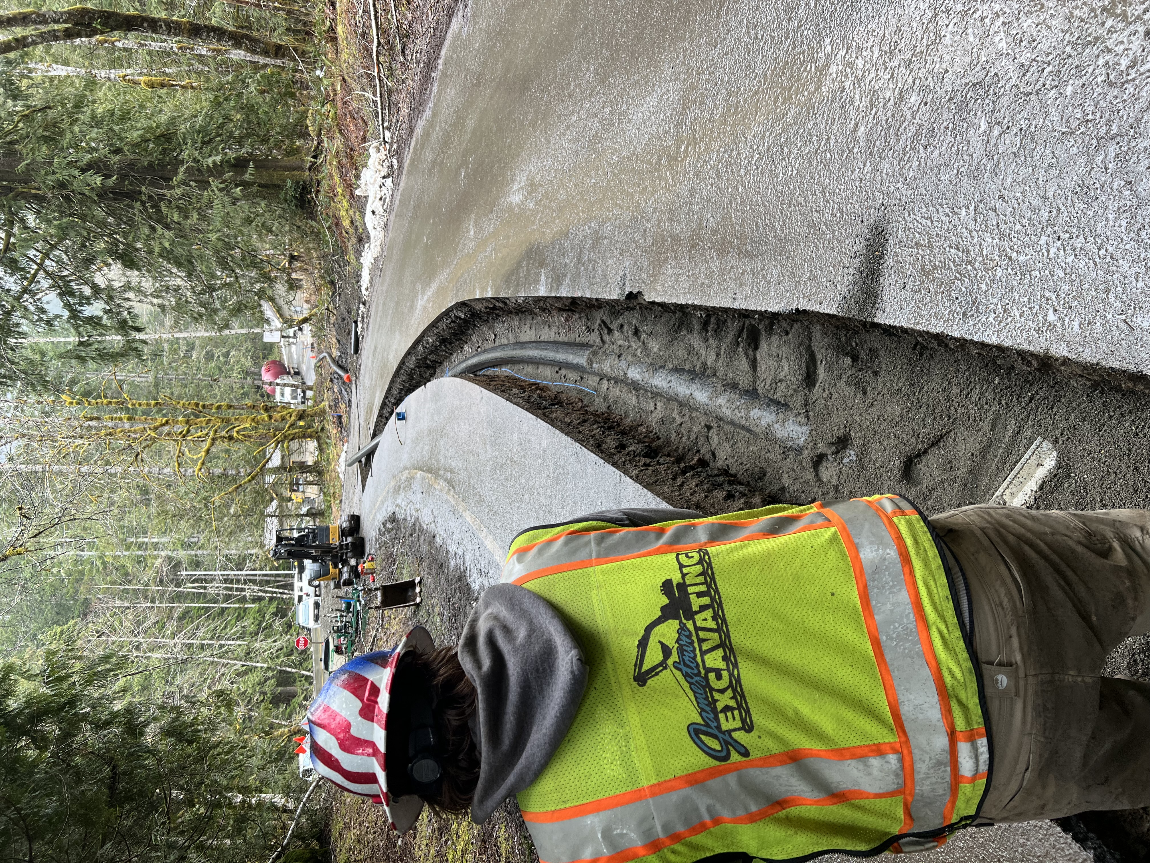 A worker in a high visibility vest stands facing a road with a trench dug in it. A pipe is laid in the bottom of the trench.