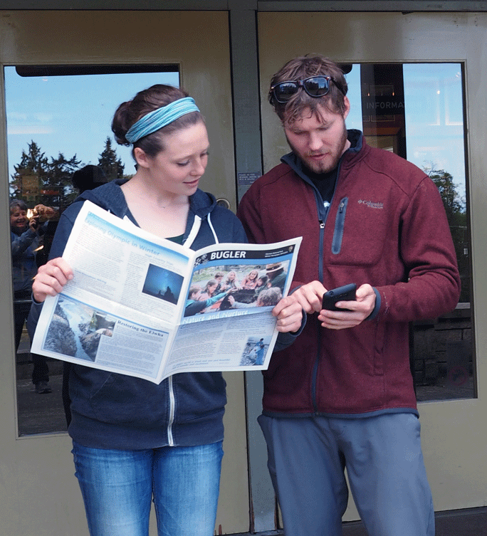 Two people standing in front of a building reading a newspaper and looking at a cell phone.