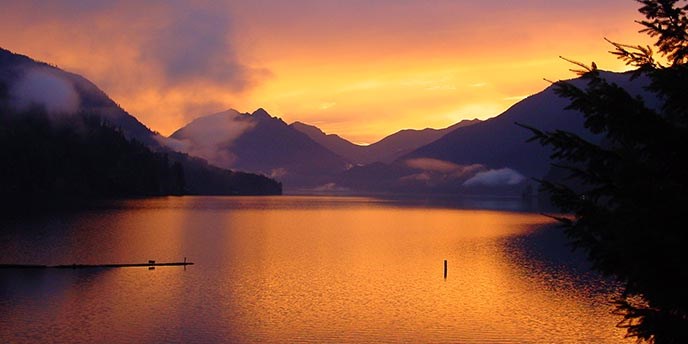 a lake at sunset reflecting shades of orange and yellow surrounded by mountains and trees