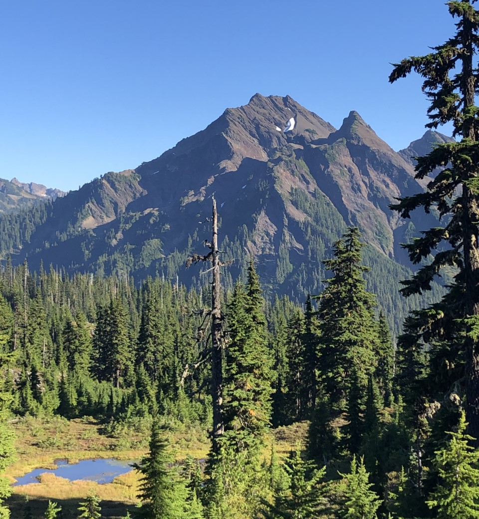 A rocky mountain peak with two tiny patches of white near the summit, and a lake in the foreground.