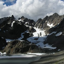 A mountain scene. A glacial lake lies in the foreground. A snowfield rises on a slope above the lake and stretches up to jagged mountain peaks. Large white puffy clouds float in the sky in the background.