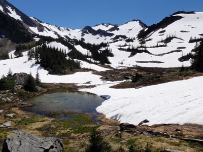 A small pond in a mountain valley. The pond has just melted out from under the snow and the rest of the valley is mostly snow-covered.