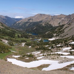 A partially snow-covered high elevation mountain basin. Small trees dot the upper basin. A small lake lies lower down and eventually the valley becomes fully forested.