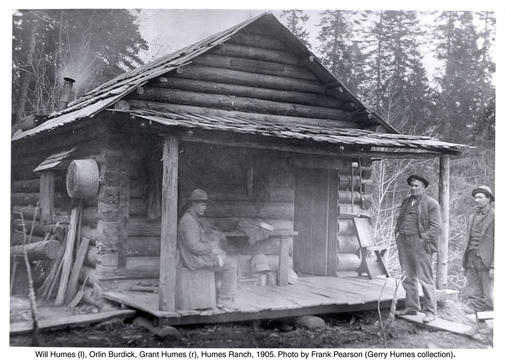 A historic black and white photo of three men outside of a small, rustic, wooden cabin. Underneath the photo reads the text: "Will Humes (l), Orlin Burdick, Grant Humes (r), Humes Ranch, 1905. Photo by Frank Pearson (Gerry Humes collection)."