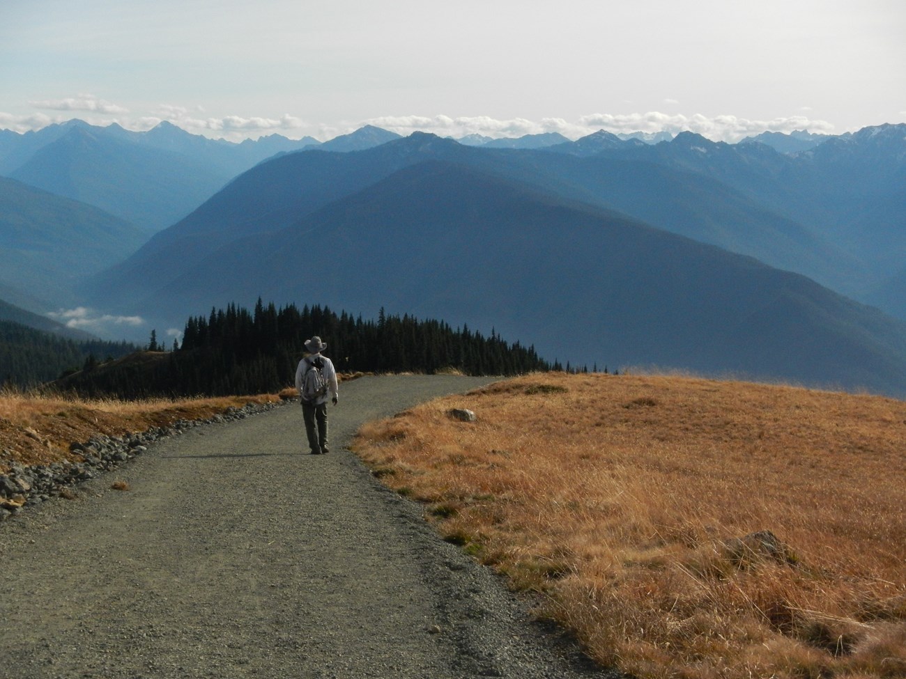A hiker on a wide gravel trail with mountains in the distance.