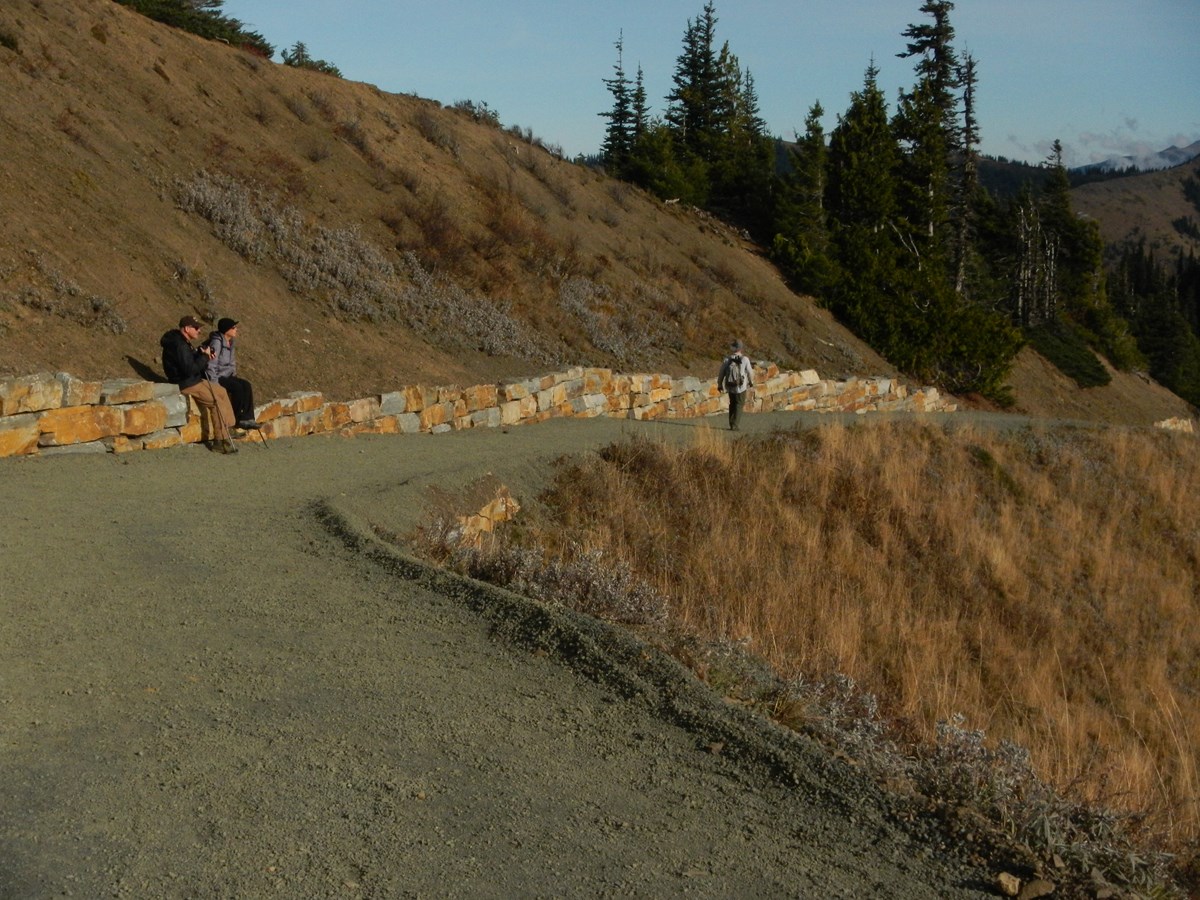 Two people sitting on a rock wall along a gravel trail in the mountains with another person visible on the trail in the distance.