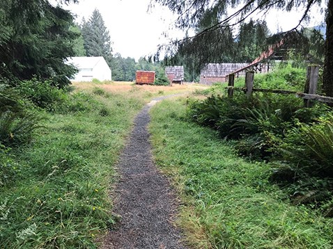 A narrow gravel trail leads through a grassy corridor along a fence. The trail and corridor eventually lead to an open grassy field. In the field sit, several old, wooden homestead buildings.