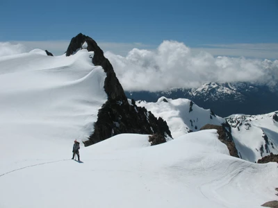 A mountaineer stands on a snowy slope high in the mountains, with lower snowcapped mountains lying below in the background.