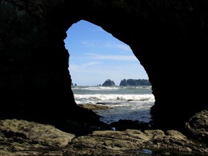 A rocky sea stack forms a natural arch. The view shows through the arch towards the ocean and additional rugged sea stacks.