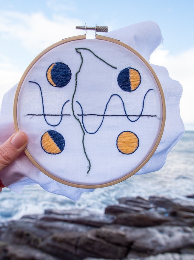 A hand holds an embroidery hoop with a design of four moons, the outline of the Olympic Coast, and an abstract tide chart. Waves crash against rocks in the blurred background.