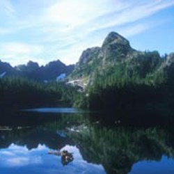A mountain peak looms behind a subalpine lake on a sunny day. The reflection of the mountain can be seen on the calm water of the lake.