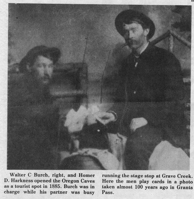 Two men sit at a small table playing cards in an old, grainy black-and-white photograph. On the right, Walter C. Burch is dressed in a suit and hat, and on the left, Homer D. Harkness sits with his hat slightly askew.