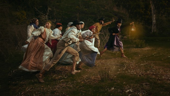 Nighttime photograph of a group of 8 African American freedom seekers in 19th century clothing, hurrying to follow a woman dressed as Harriet Tubman and holding up a lantern.