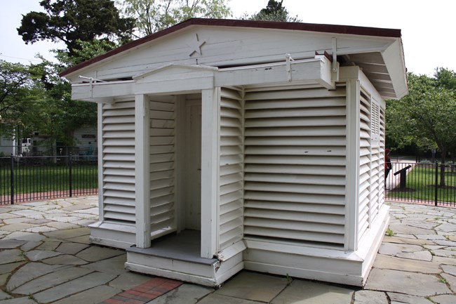 A small white wooden building with louvered exterior walls in the center of a flagstone plaza with green trees in the background.