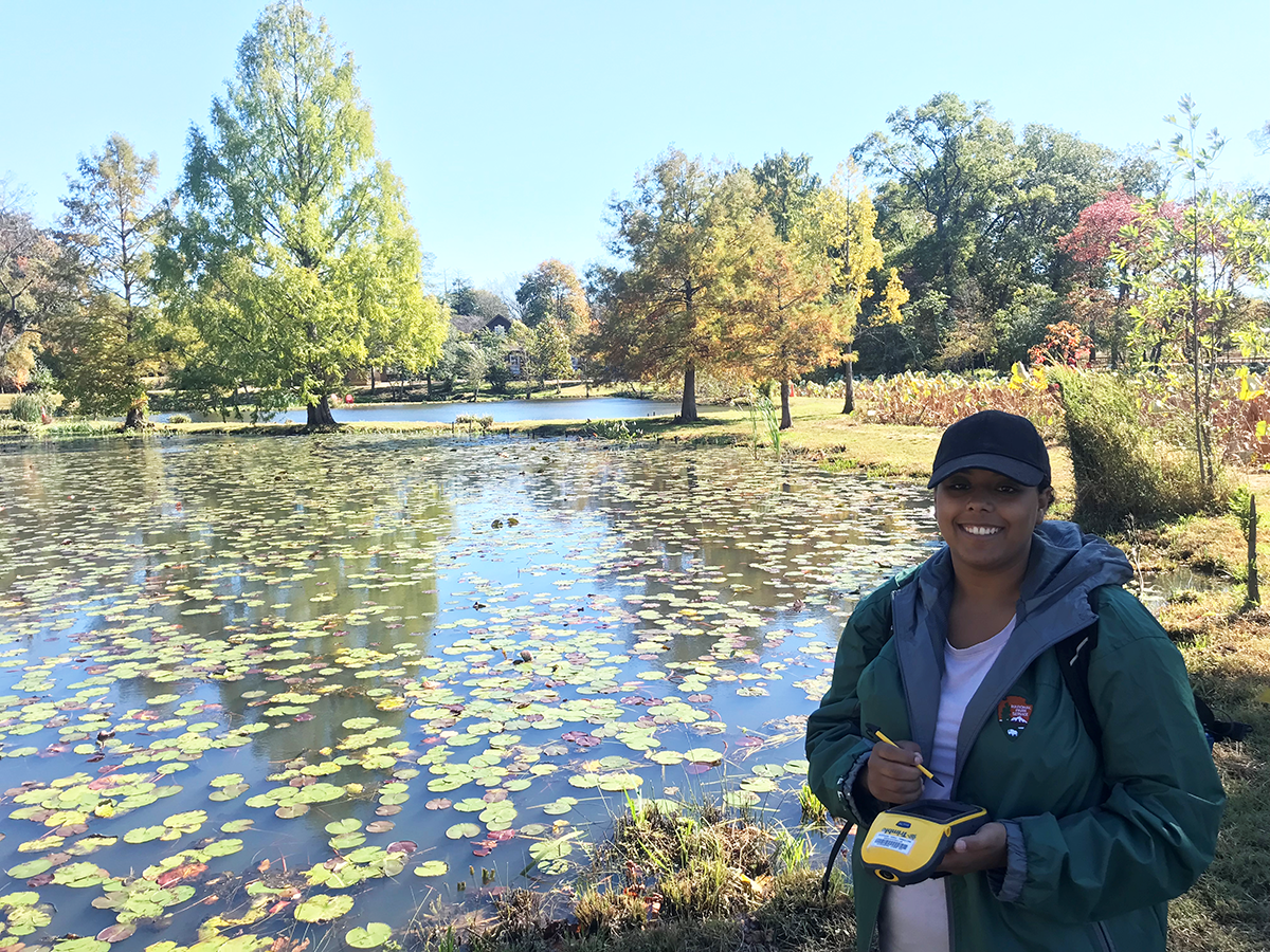 Geolocating at Kenilworth Woman stands holding a yellow digital unit with lily pad covered pond in background