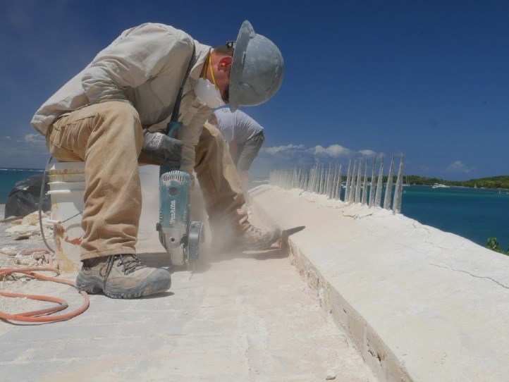 A person wearing a hard hat, protective glasses, gloves, and a mask, uses a tool to cut concrete.