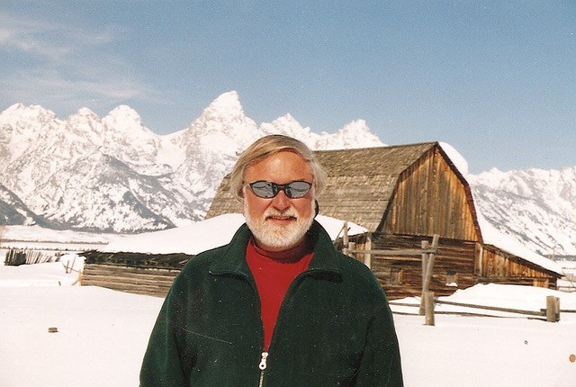 Harrison Goodall A man with white hair and a white beard smiles standing in front of a historic barn with snowy mountains in the distance.