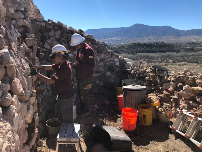Two workers wearing maroon shirts and hard hats replace mortar between large stones of a prehistoric home