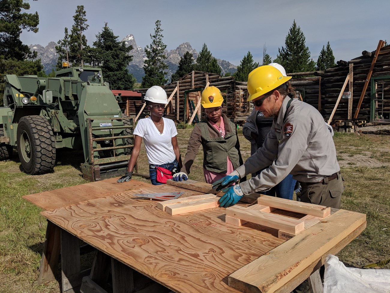 Four people wearing hard hats and tool belts work on a piece of wood in front of a large mountain range.