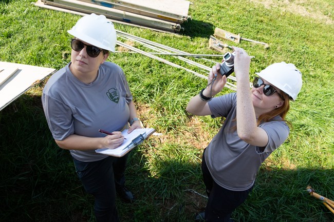 Two interns wearing NPS tshirts and hard hats look up. One of the interns has a clipboard and the other has a camera.