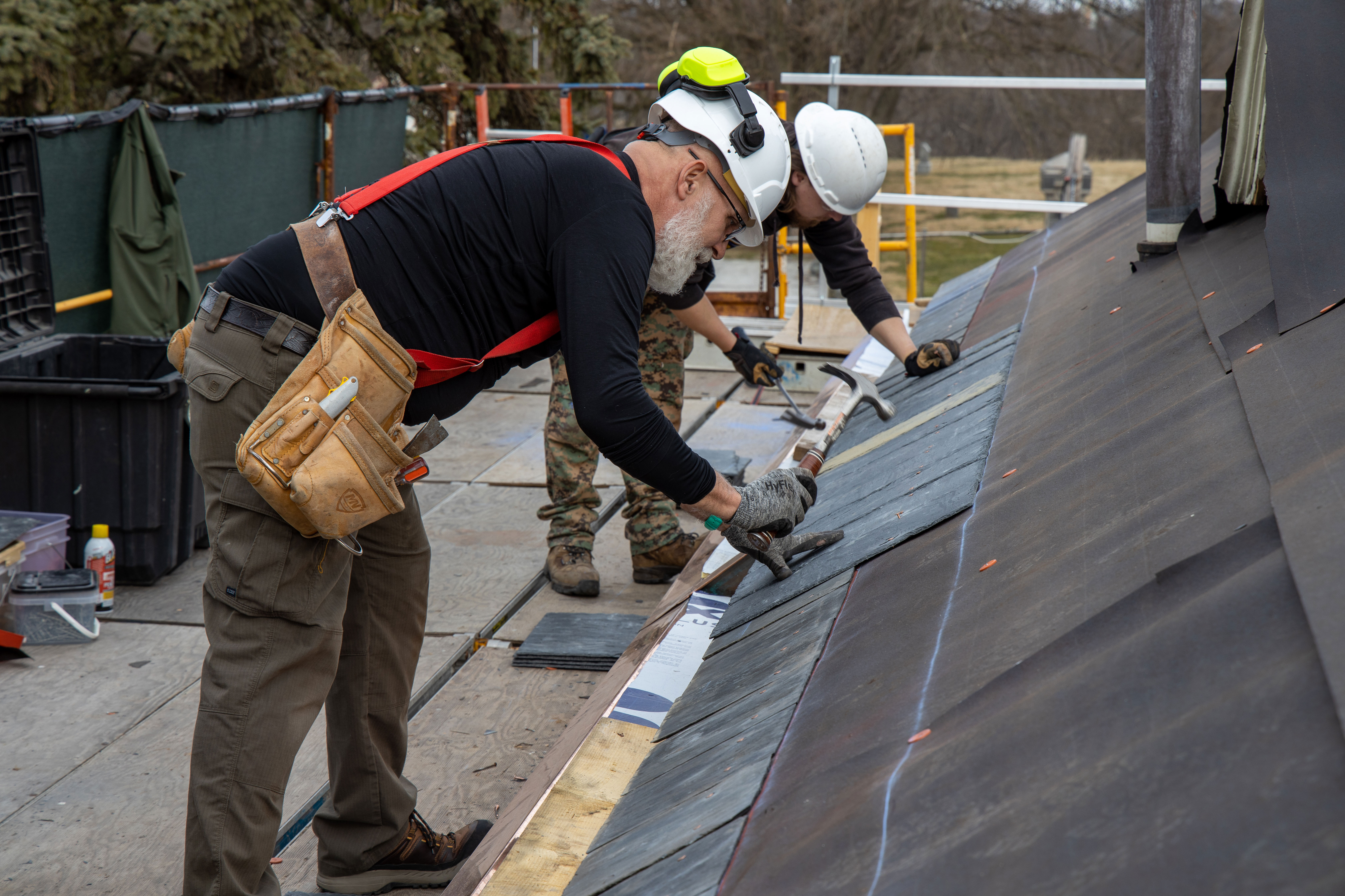 Roofing Project Two workers wearing hard hats and safety gear nail slate tiles on a roof.