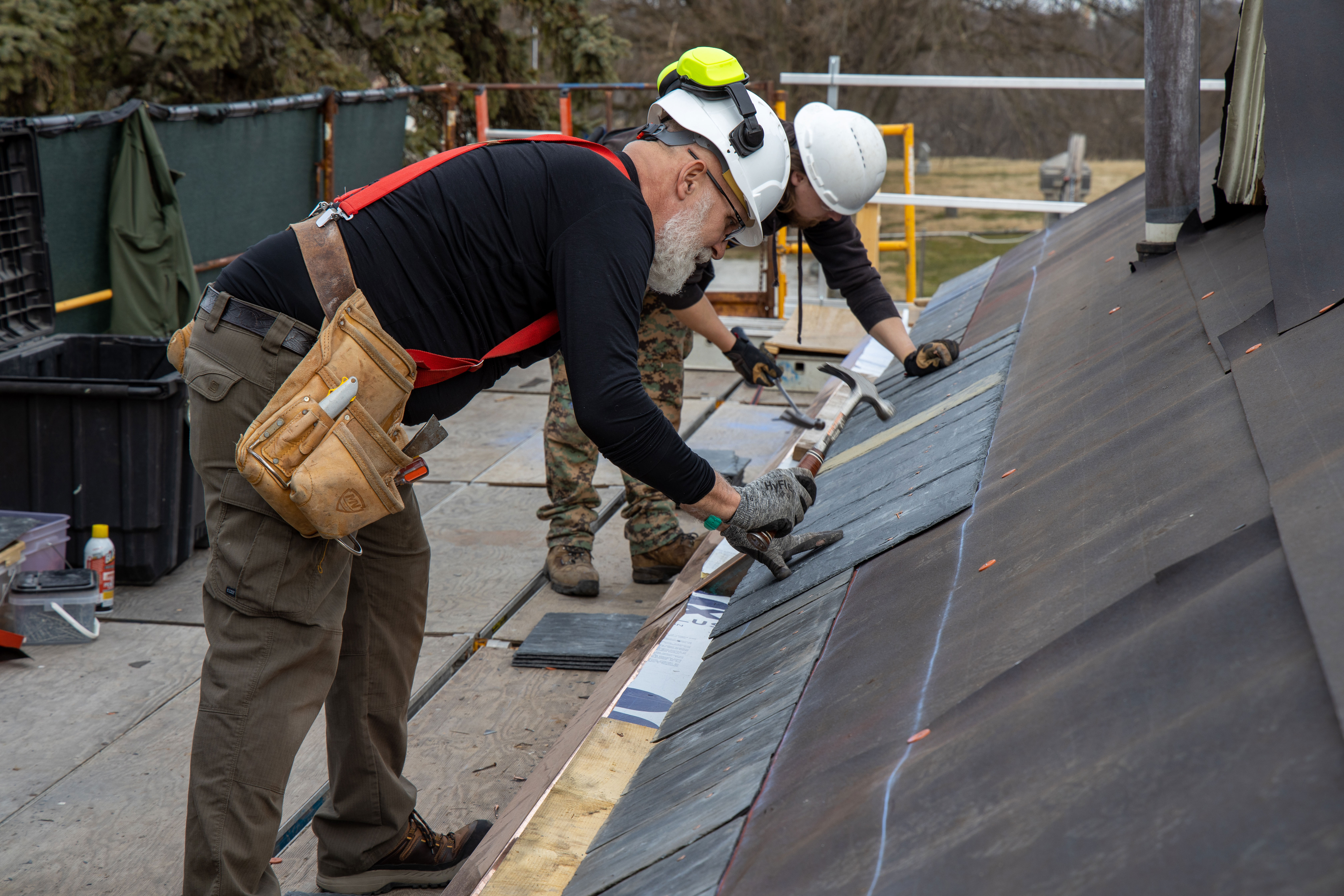 Two workers wearing hard hats and safety gear nail slate tiles on a roof.